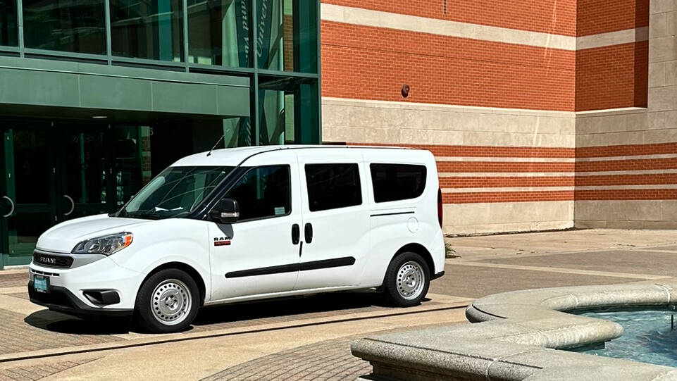 White van parked near a building with large glass windows and red brick walls. A fountain is on the right.